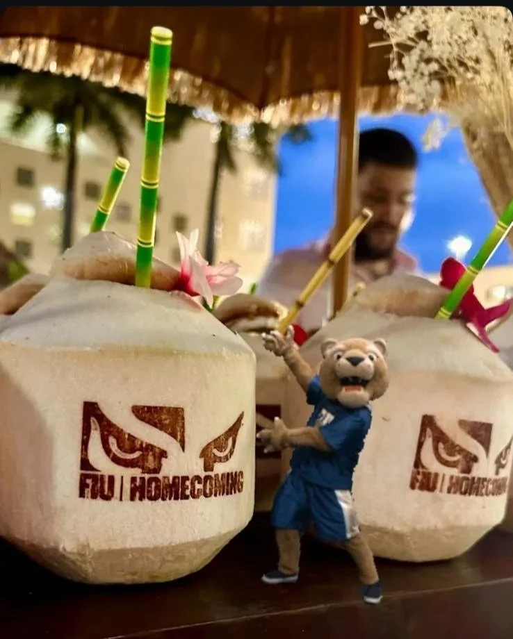Two coconut drinks with straws and flowers on a table, a person in the background, and a lion mascot in front of the coconuts, all at an outdoor setting.