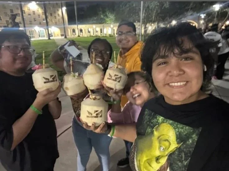 Some people are smiling and holding logo-printed coconuts with straws, outdoors at night.