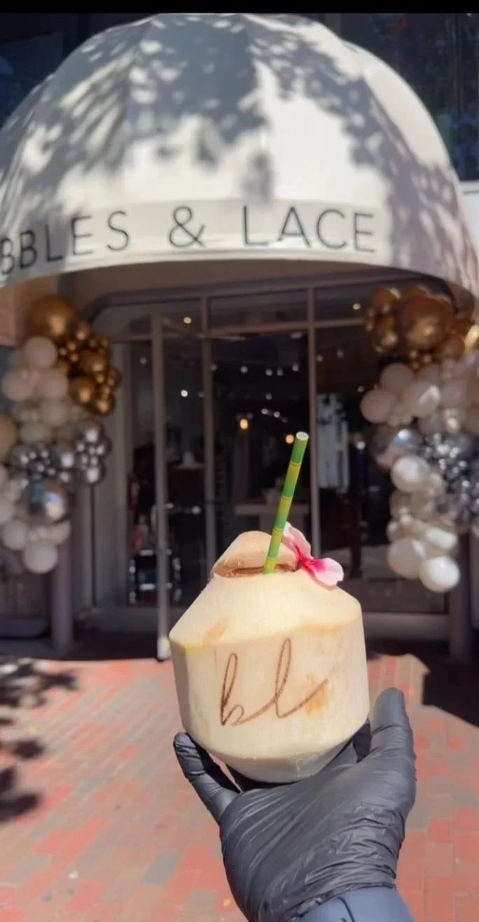 Person holding a fresh young logo coconut with a straw, in front of a store with a white awning reading "GOBBLES & LACE" decorated with balloons.