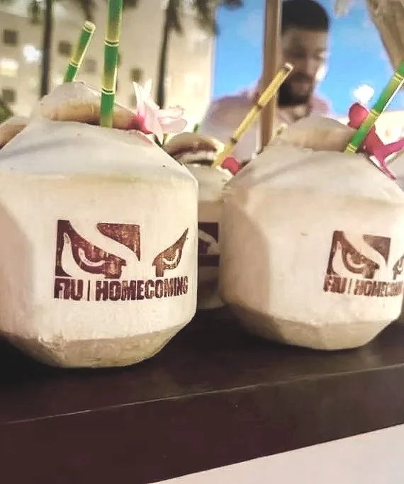 Two coconuts with straws and flowers, each featuring a logo that reads 'FUI HOMECOMING' with an owl's eyes and beak design.