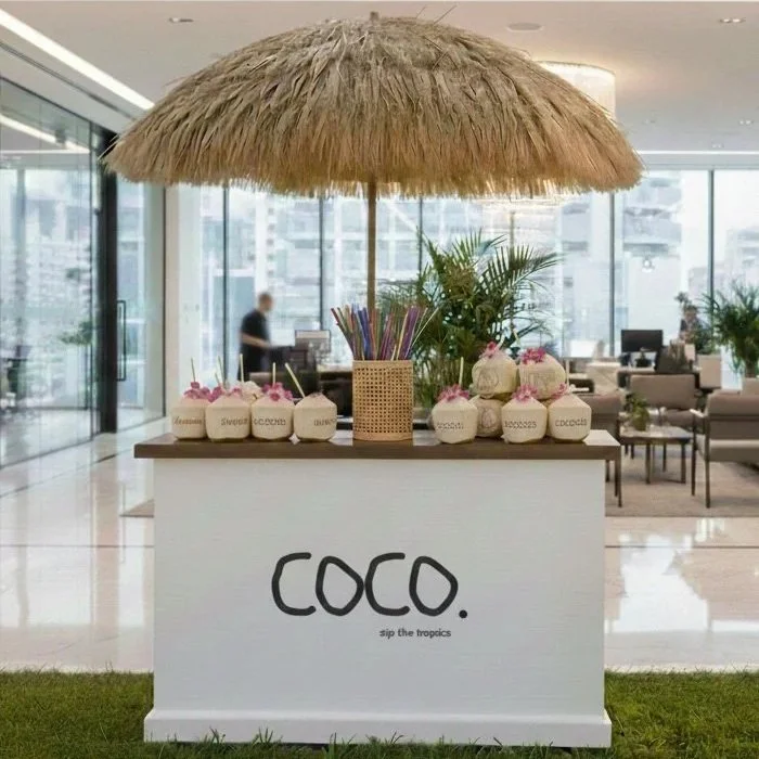A tropical-themed coconut stand with a thatched umbrella, decorated coconuts, colorful straws, and a sign that reads 'COCO. sip the tropics' in a bright, modern indoor setting.