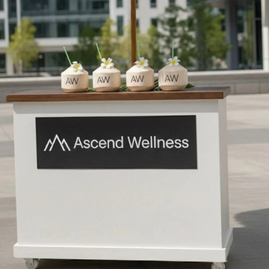 A wellness event registration desk with four coconut drinks decorated with flowers and 'AW' stir sticks, and a sign that reads 'Ascend Wellness' with a mountain logo.