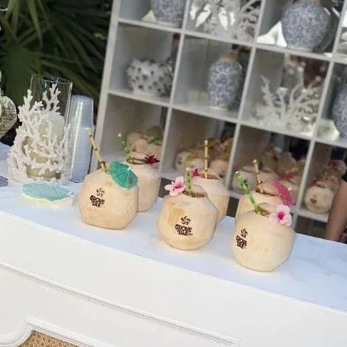 Four coconut drinks with straws and small pink and green flowers on a white table, with decorative items and shelves in the background.