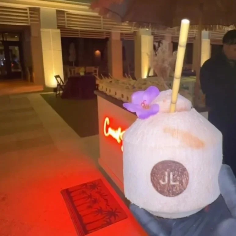 A person holding a coconut drink with a straw, an orchid flower, and a purple flower on top, in a bar or restaurant with a welcoming ambiance, warm lighting, and a red carpet.