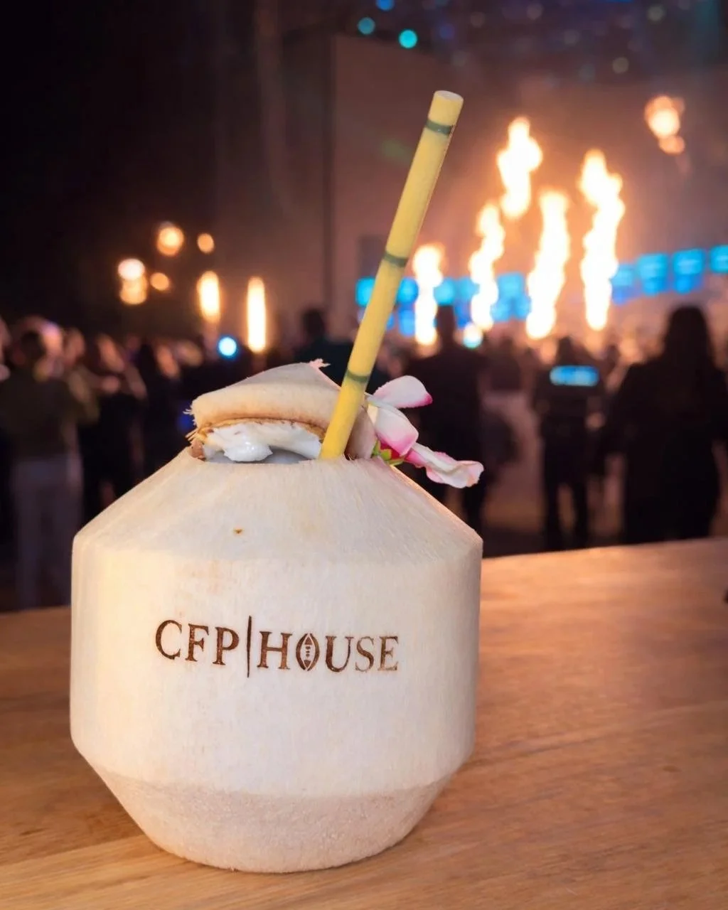 A fresh coconut with a straw and flower on a wooden table in front of a concert stage with flames and blue lights.