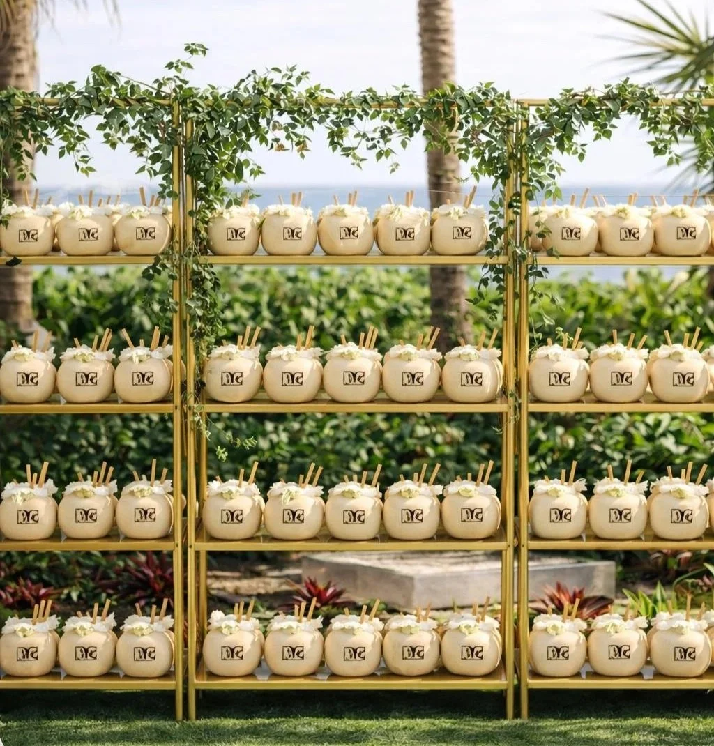 Multiple white coconut drinks with black logos on a yellow metal shelf outdoors, decorated with green foliage and in the background palm trees and a body of water.