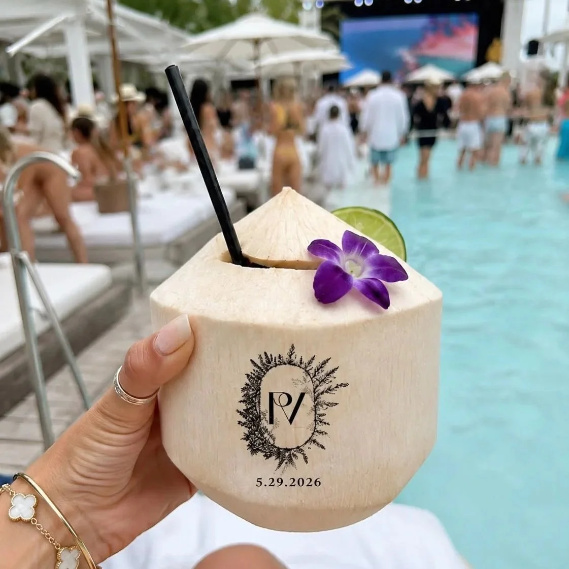 Hand holding a fresh coconut drink with a black straw, lime wedge, and purple flower at a poolside party with people and umbrellas in the background.