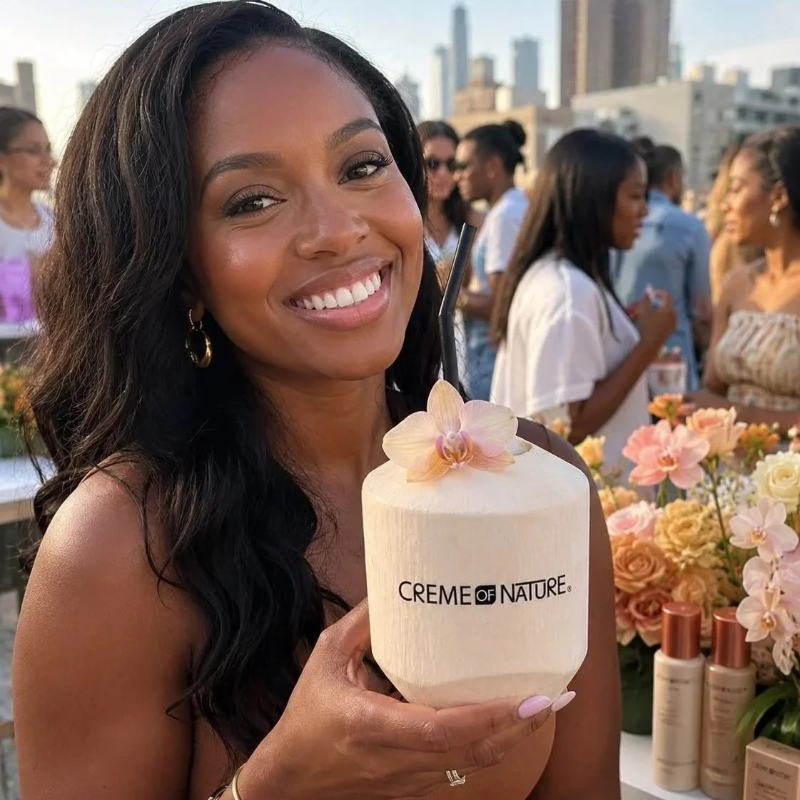 A woman with dark wavy hair smiling and holding a coconut-shaped drink with a straw and an orchid decoration, at an outdoor event with people and flowers in the background.