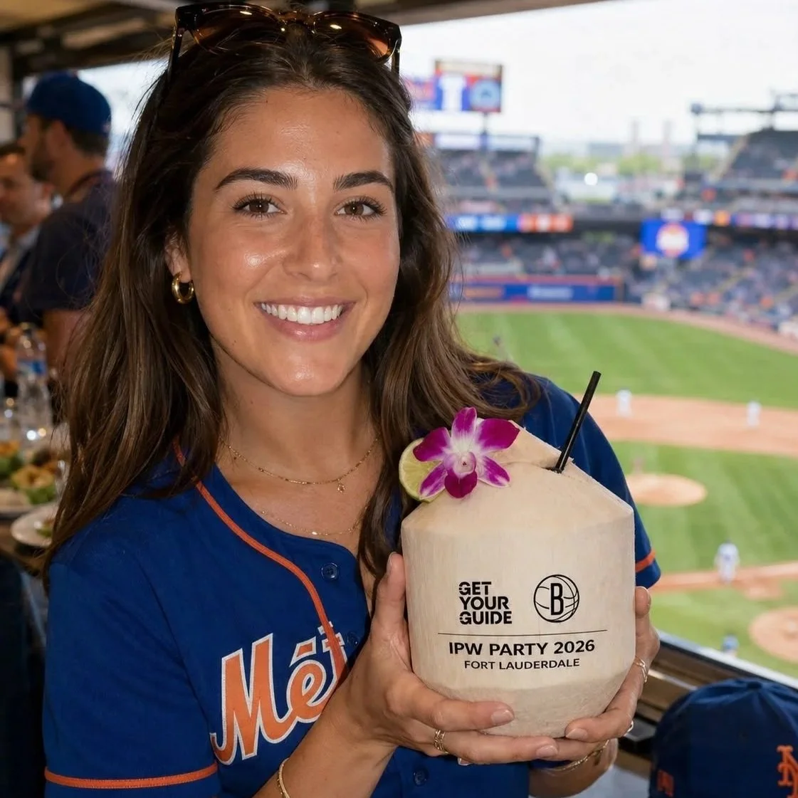 A smiling woman wearing a blue baseball jersey holding a coconut beverage decorated with an orchid flower at a baseball stadium.