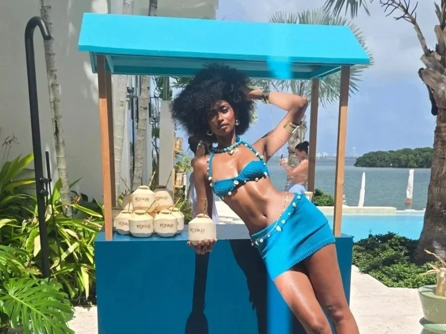 A woman in a blue bikini and matching skirt standing next to a blue stand with coconuts and a beverage, outdoors near a pool with water and trees in the background.