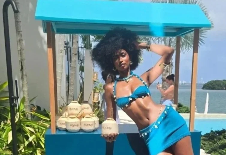 A woman in a blue bikini and skirt posing by a beachside kiosk with coconut drinks, with a body of water and a woman in the background.