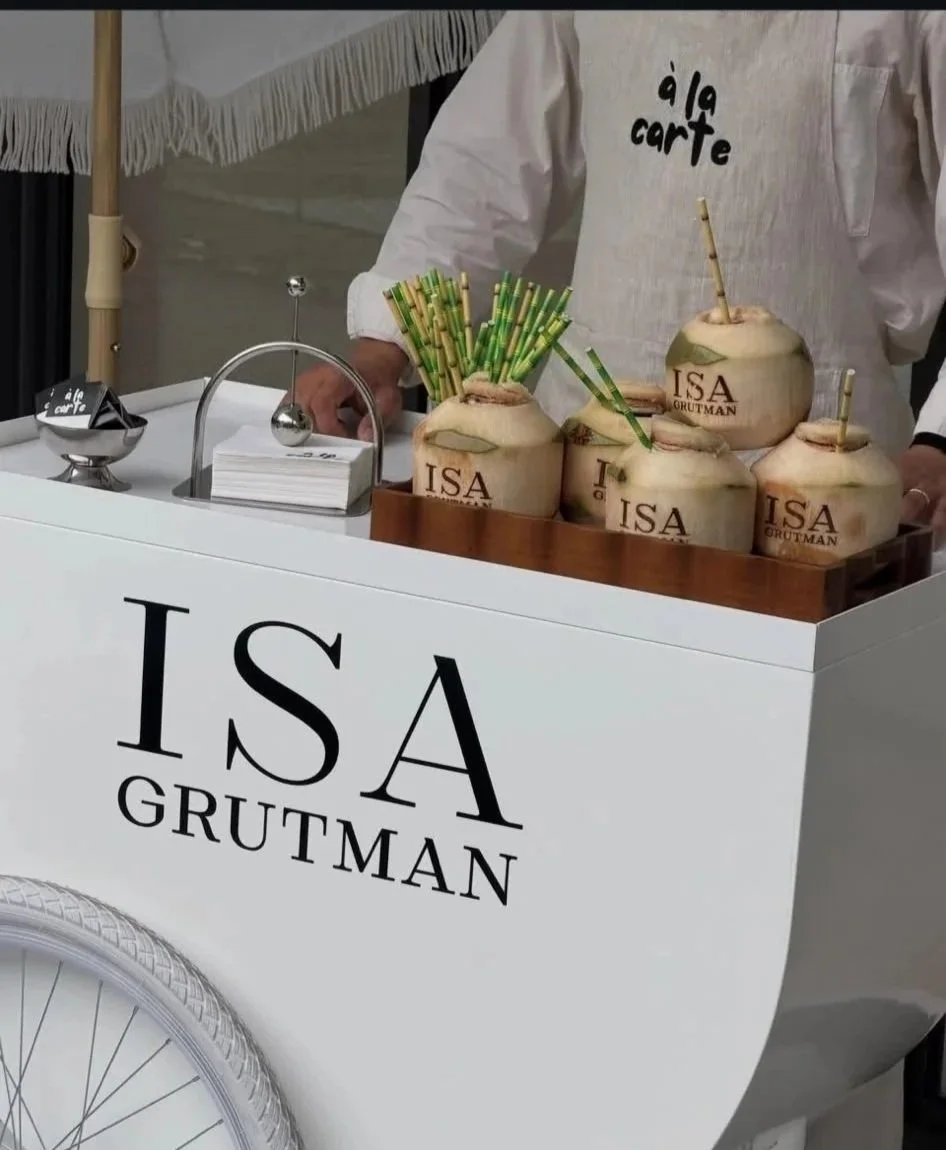 A white cart with the words 'ISA GRUTMAN' on the front. On top of the cart, there are several coconuts with straws and the logo 'ISA GRUTMAN' engraved on them, serving drinks. A person wearing a white apron with 
