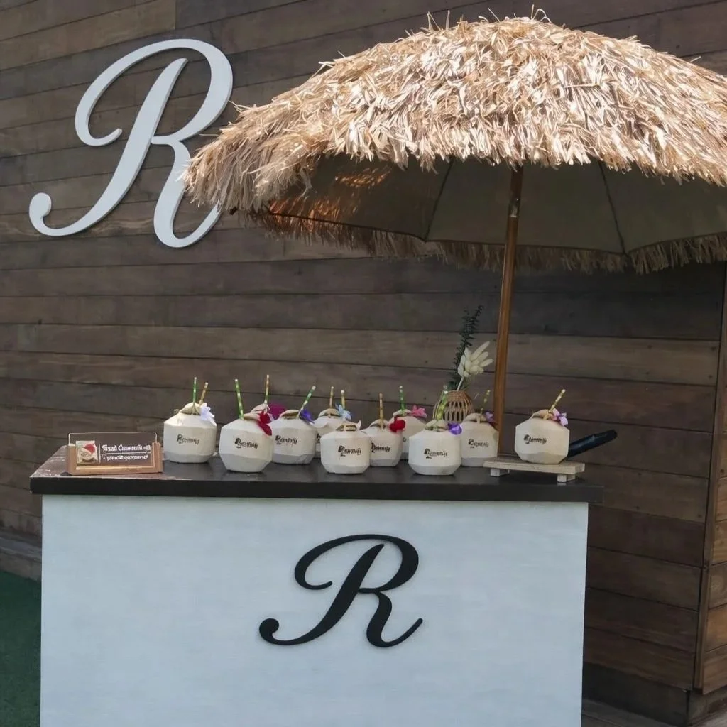 A drink station with seven coconut beverages decorated with flowers and paper umbrellas, placed on a counter with a large letter 'R' on the front. An umbrella shades the station, and a wood wall with a large 'R' is in the background.