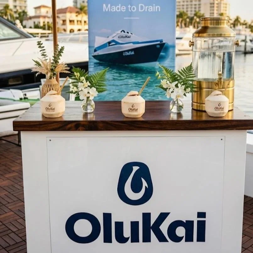 A promotional display featuring a boat with the text "Made to Drain" on a banner. The station has small containers with the Olukai logo, floral arrangements, and a large lantern, all set on a wooden table with a marina and boats in the background.