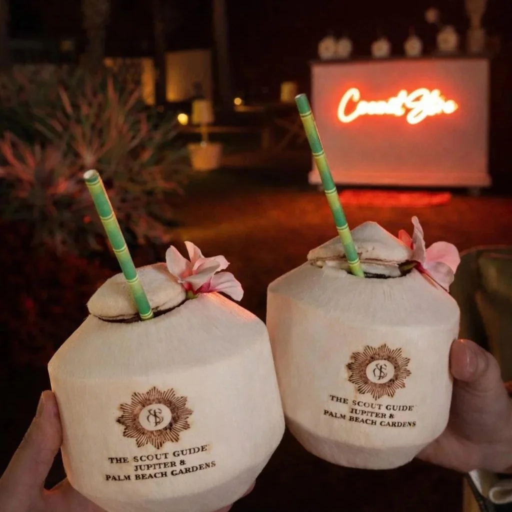Two beige coconuts with pink flowers and green-striped straws held by hands, in a tropical setting at night with a neon sign in the background that reads 'Come Bar'.