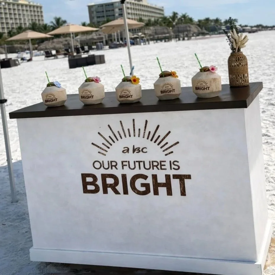 Beachside bar with five coconuts with drinks and flowers on top, next to a white stand with the text 'a bc OUR FUTURE IS BRIGHT' and a sun illustration, in front of a sandy beach with umbrellas and buildings in the background.