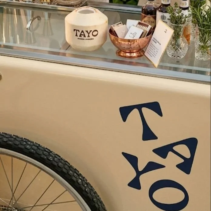 Close-up of a beige-colored surface with a large blue 'TAYO' logo and a bicycle wheel in the lower left corner. The background shows a glass display case with a white container labeled 'TAYO,' a copper bowl with small white boxes inside, bottles, and