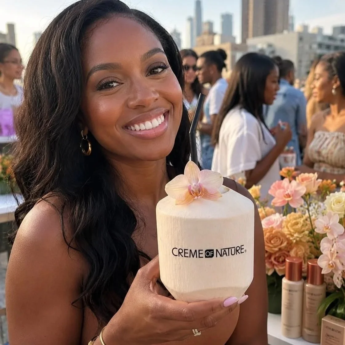 A smiling woman with dark wavy hair holds a coconut-shaped container with a flower on top, standing at an outdoor event with people and city buildings in the background.
