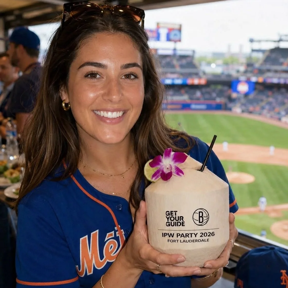 A smiling woman in a Mets jersey holding a coconut drink with a pink flower and lime wedge during a baseball game at a stadium.