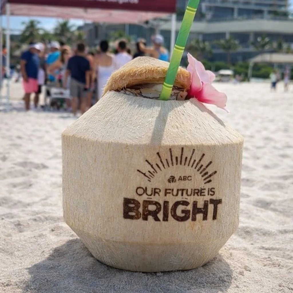 A coconut drink with a straw and a pink flower on a sandy beach, with a crowd of people and buildings in the background.