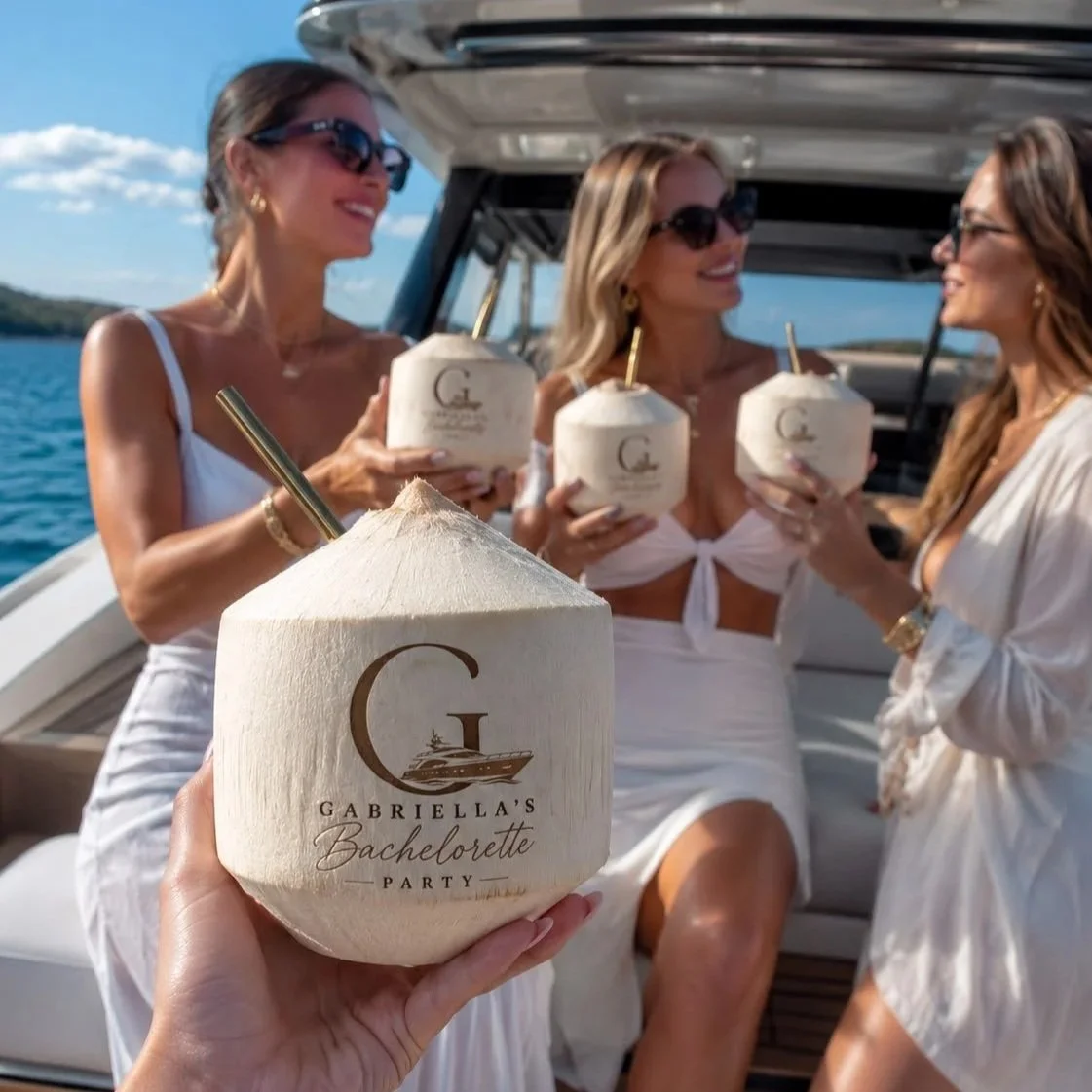 Four women wearing white dresses and sunglasses on a boat, holding coconuts with straws, celebrating a bachelorette party.