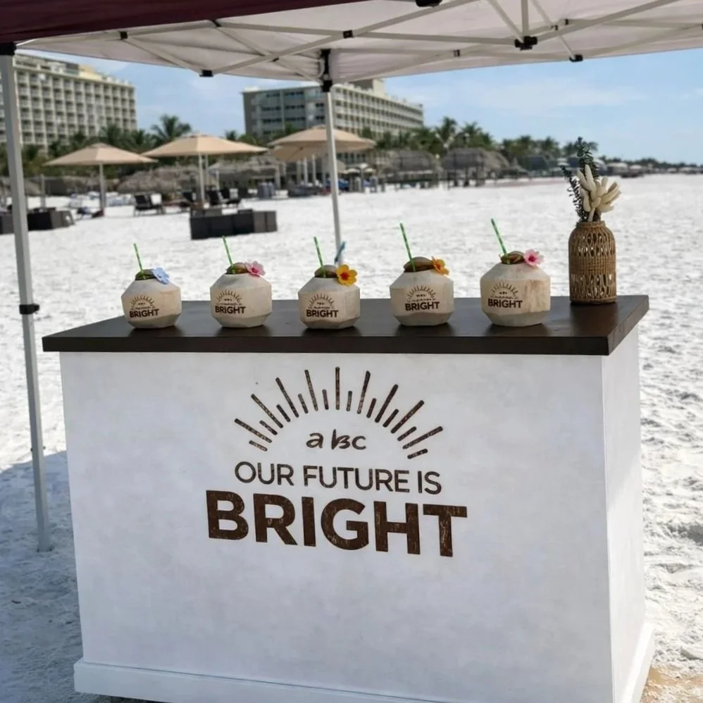 Beachside booth with four coconut drinks decorated with small flowers and straws, and a vase with white flowers, set against a backdrop of beach umbrellas and buildings on a sandy beach.