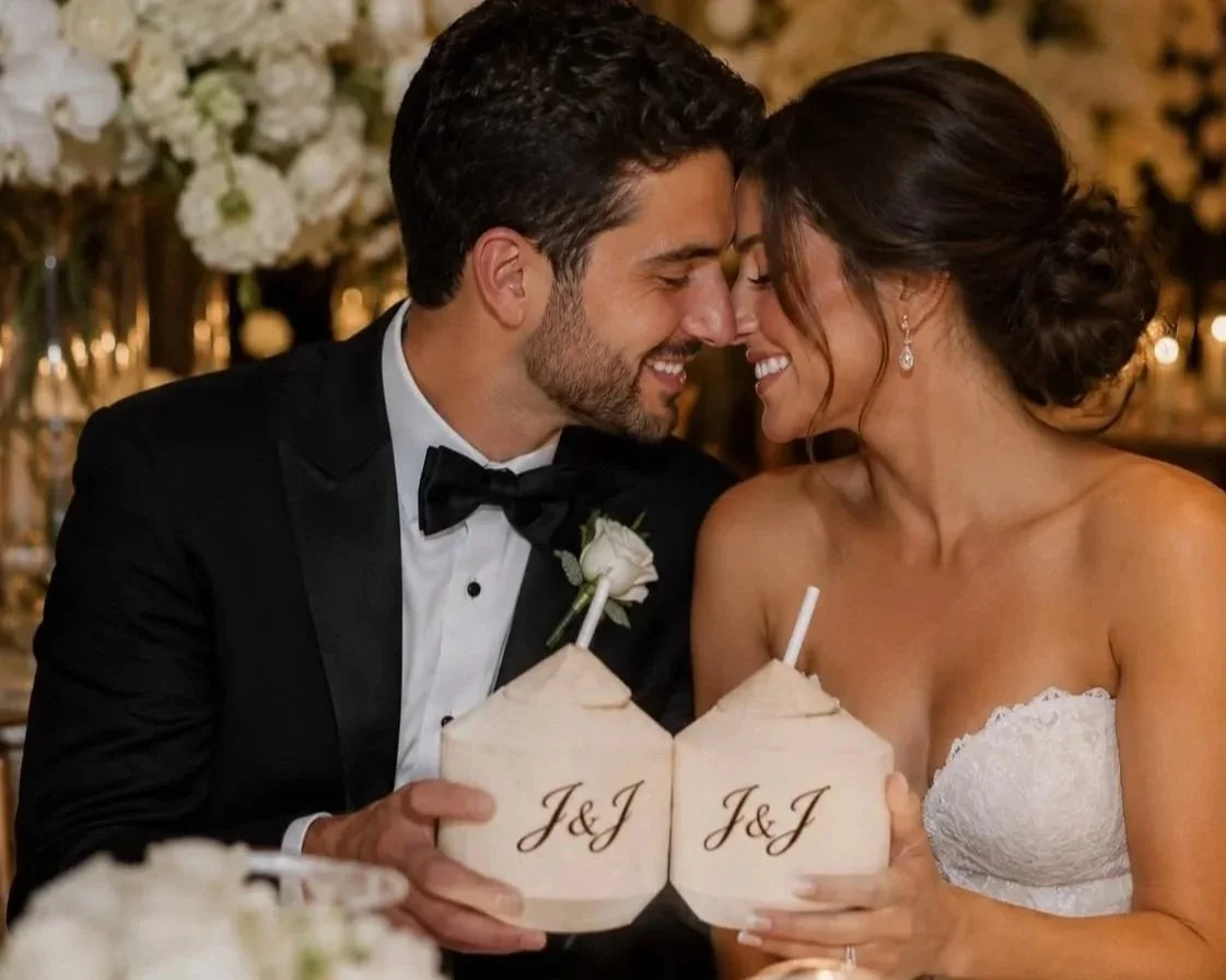 A newlywed couple touching heads and smiling at each other while holding personalized coconut drinks with 'J & J' written on them during their wedding celebration.