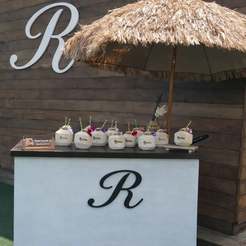 A beverage station with a straw-covered umbrella, six white coconuts with colorful straws and flowers, and a small card on a black and white stand. The station has a large black letter 'R' on the front, set on a wooden backdrop with a 'Welcome' mat f