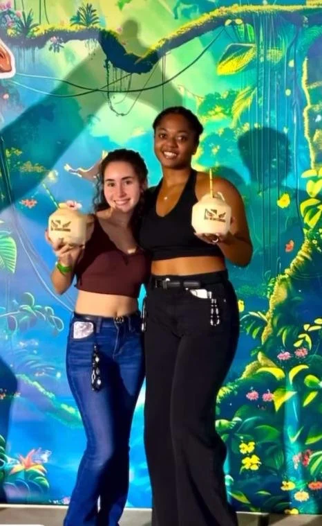 Two young women standing side by side in front of a colorful jungle-themed mural, each holding a coconut with a straw and a logo on it, smiling at the camera.
