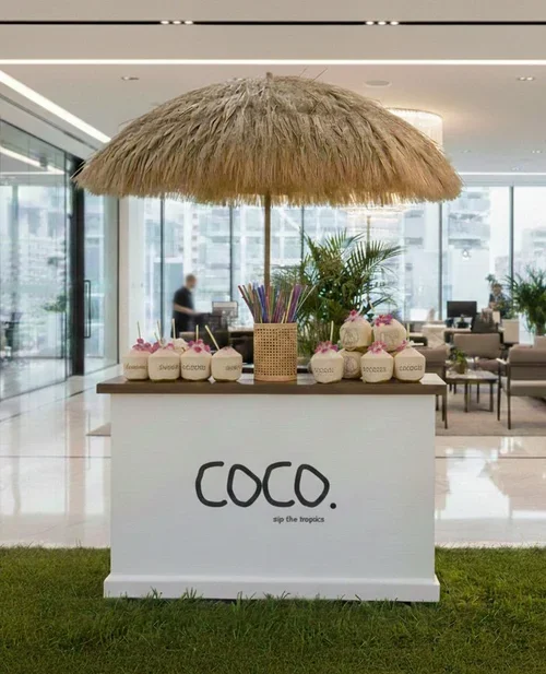 A tropical-themed coconut stand with a thatched roof, decorated with coconuts, straws, and flowers, located inside a modern building with glass windows and seating areas.