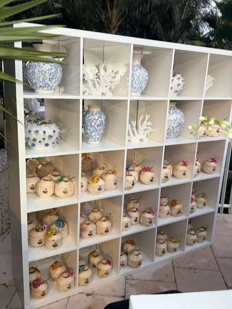 A white cubical shelving unit with various decorative items. The top row contains blue and white vases and white coral decorations. The middle and bottom rows hold beige pots with tiny colored decorations and rounded handles, arranged neatly.