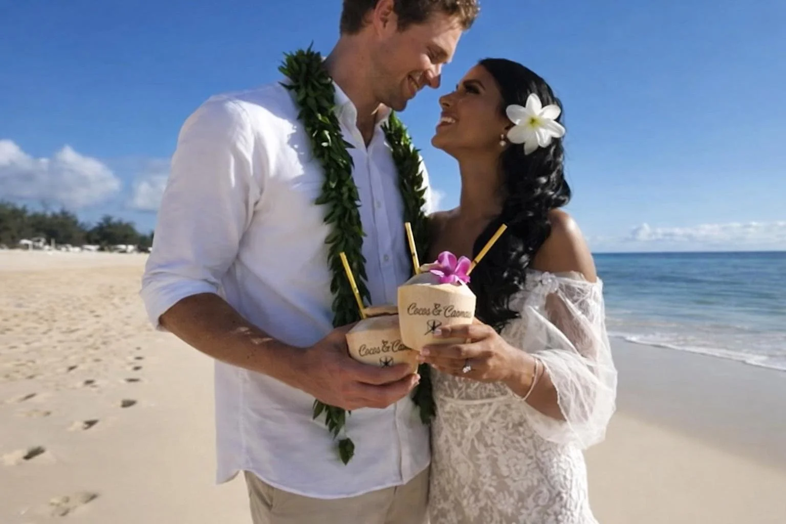 A newlywed couple on the beach holding coconuts with straws and flowers, smiling at each other with the ocean and sky in the background.