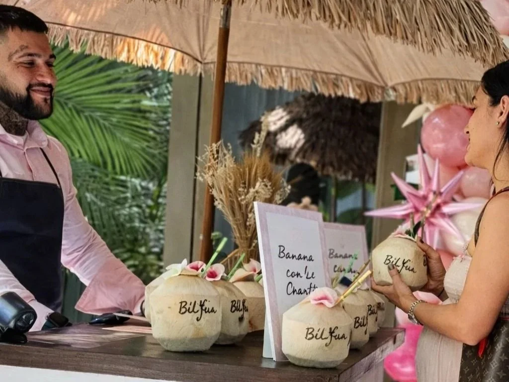 A woman purchasing fruit from a man at a tropical-themed market stall, with signs and decorative palm and flower arrangements.