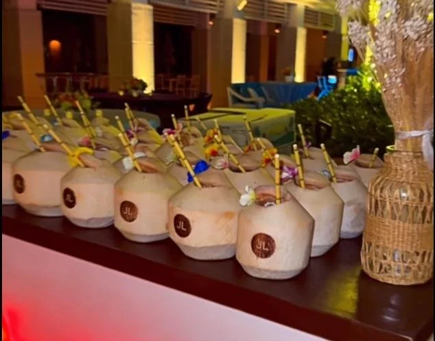 Multiple coconuts with straws and flowers on a table at an outdoor or tropical setting.