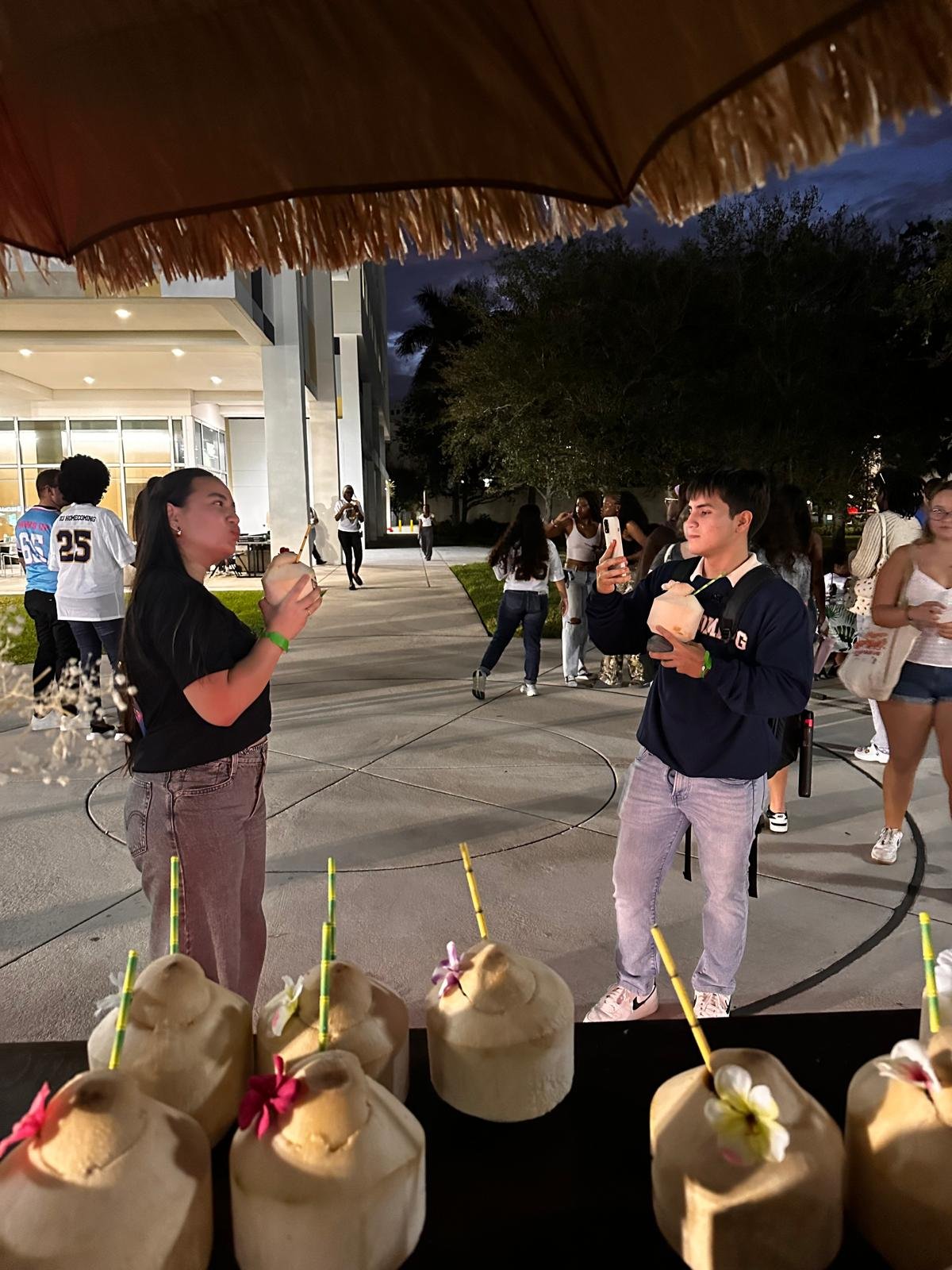 People gathered outside at night, two individuals in the foreground holding coconuts, surrounded by a group of people socializing at Florida International University, with a table of logo coconuts decorated with flowers in the foreground.