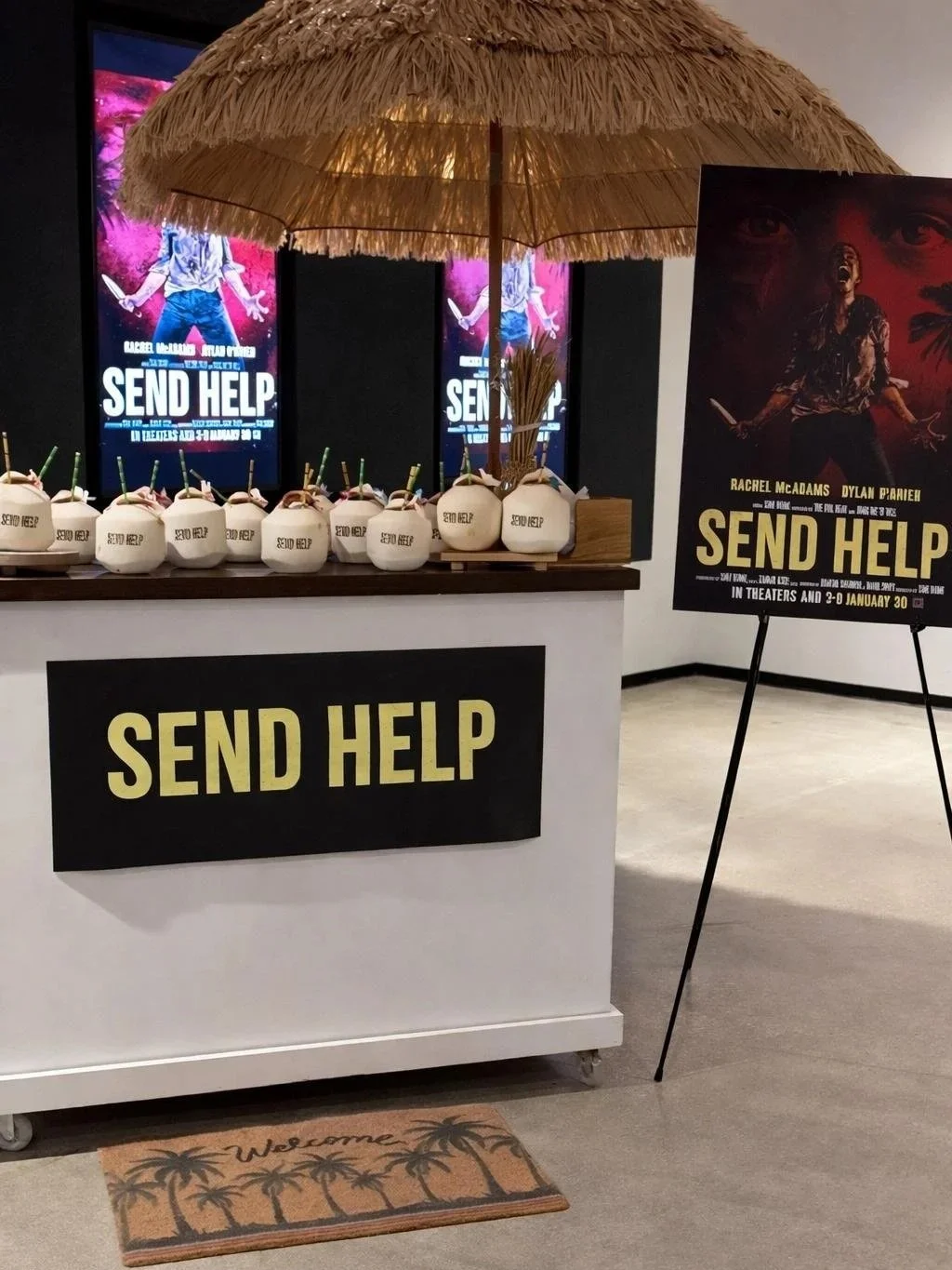 A promotional display for the movie 'Send Help' featuring a large poster with a woman and weapons, multiple coconuts with straws on a counter, a decorative umbrella, and promotional signs.