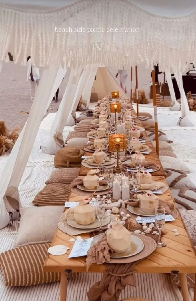 Long wooden table set for a beachside picnic celebration with plates, glasses, candles, branded coconuts and decorative elements, shaded by a fringed canopy, with cushions and rugs on the sand surrounding it.