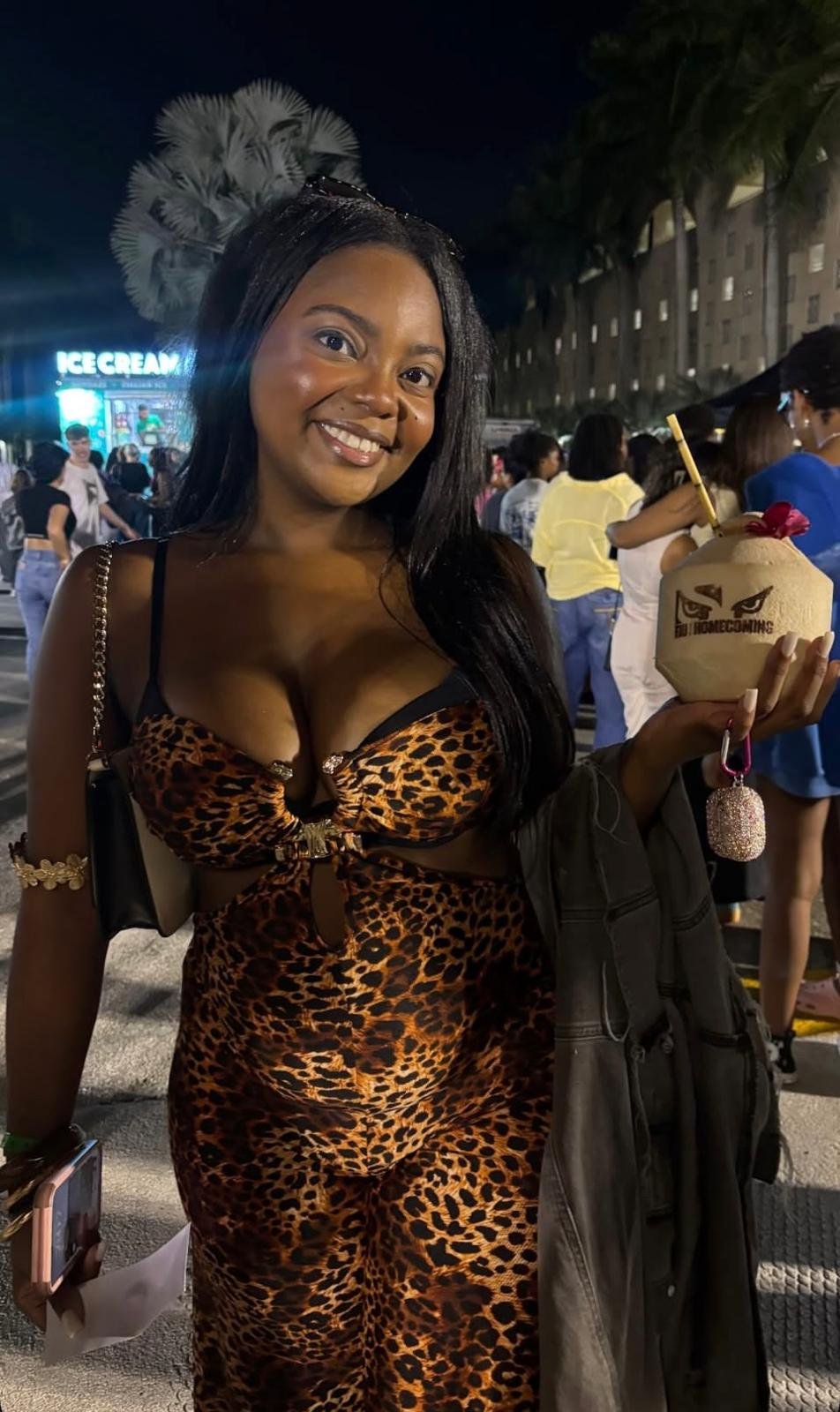 A woman in a leopard print dress, smiling at night, holding a branded coconut drink.