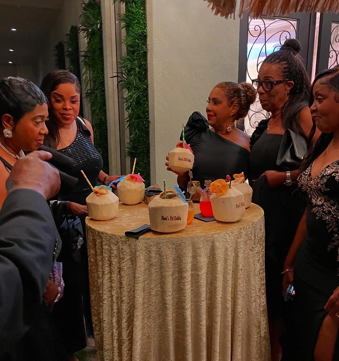 Women gathered around a table with custom-branded coconuts and drinks, celebrating at a social event, dressed in elegant black attire.
