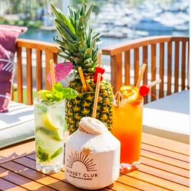 Three colorful tropical drinks with garnishes and a coconut with a straw, set on a wooden table outdoors near a marina.