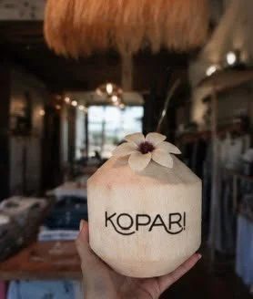 A hand holding a coconut with a flower on top and the word 'KOPAR!' printed on it in a rustic cafe setting.