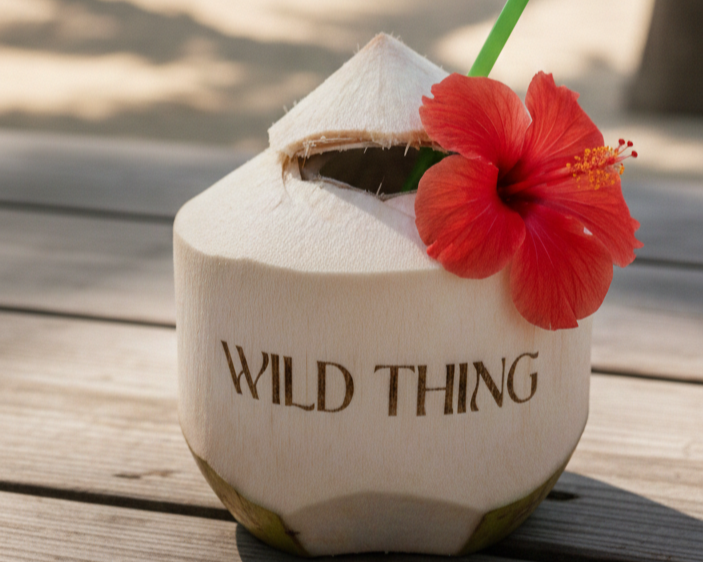 A coconut with a straw and a red hibiscus flower on top, with the words 'Wild Thing' carved into the coconut shell, placed on a wooden surface.