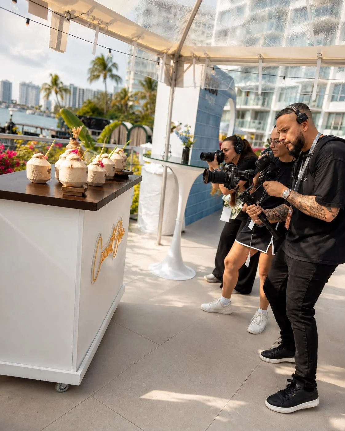 Three photographers filming a tropical-themed coconut drink stand on an outdoor patio with high-rise buildings in the background.