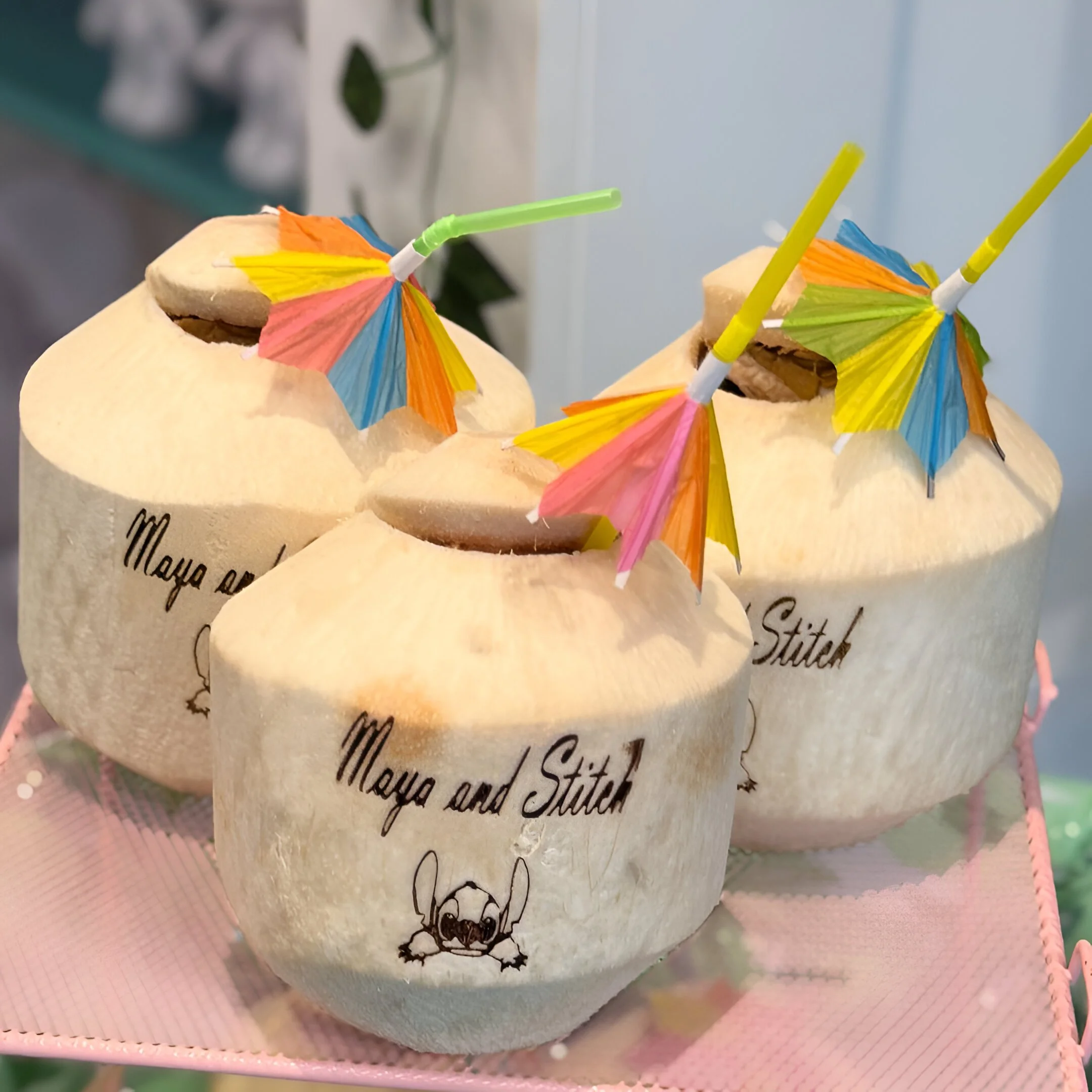 Three fresh coconuts with colorful paper umbrellas and straws on a pink tray.