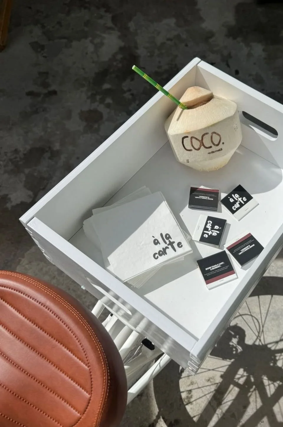 A white tray holding paper napkins, business cards, and a coconut drink with a green straw. The coconut has the word 'COCO' written on it. There is a brown leather seat nearby, and the shadow of a bicycle is cast on the ground.