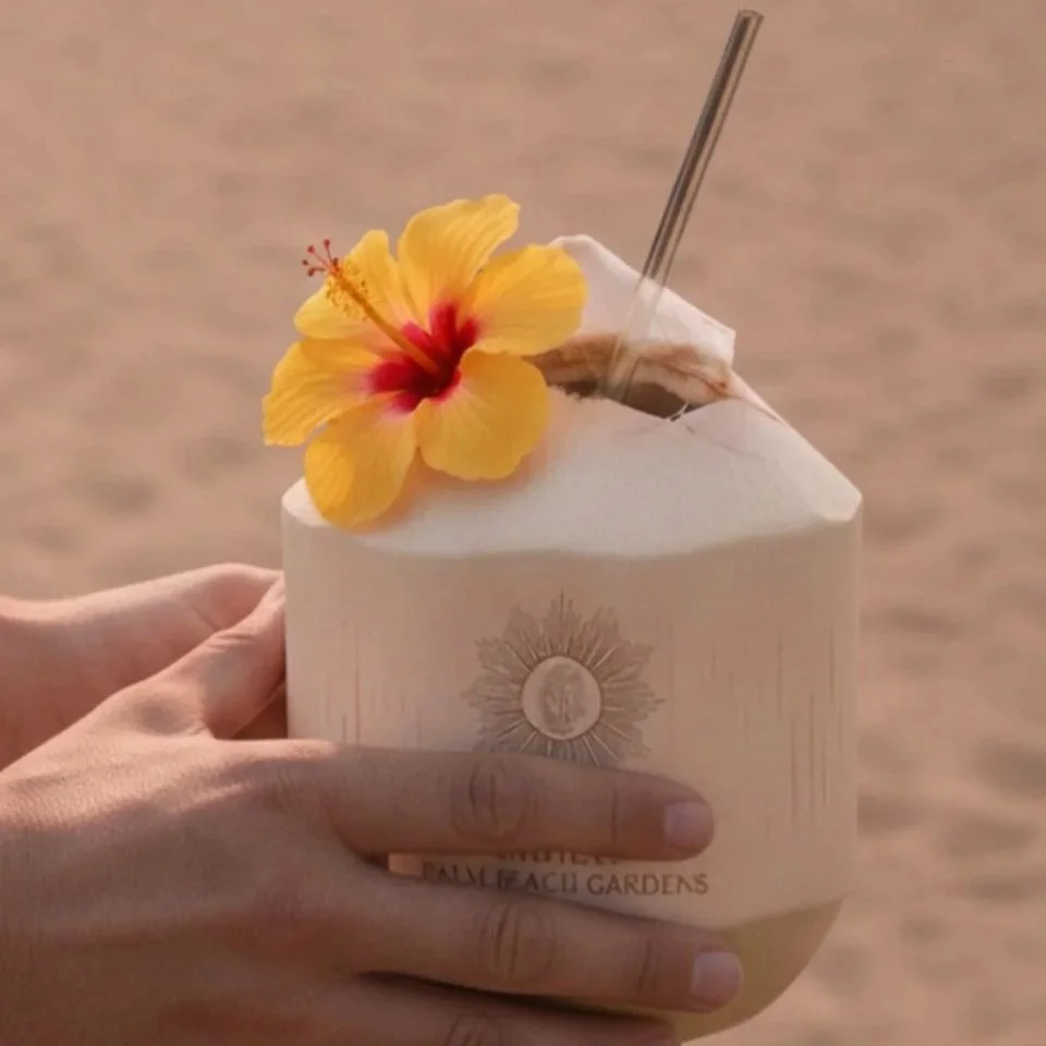 A hand holding a coconut drink with a straw, a yellow hibiscus flower, and a slice of coconut on top, in front of a sandy beach background.