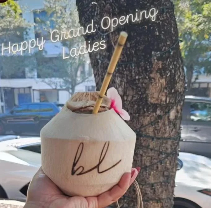 A coconut with a logo, a straw, and a pink flower on top, held by a person's hand in front of a tree.
