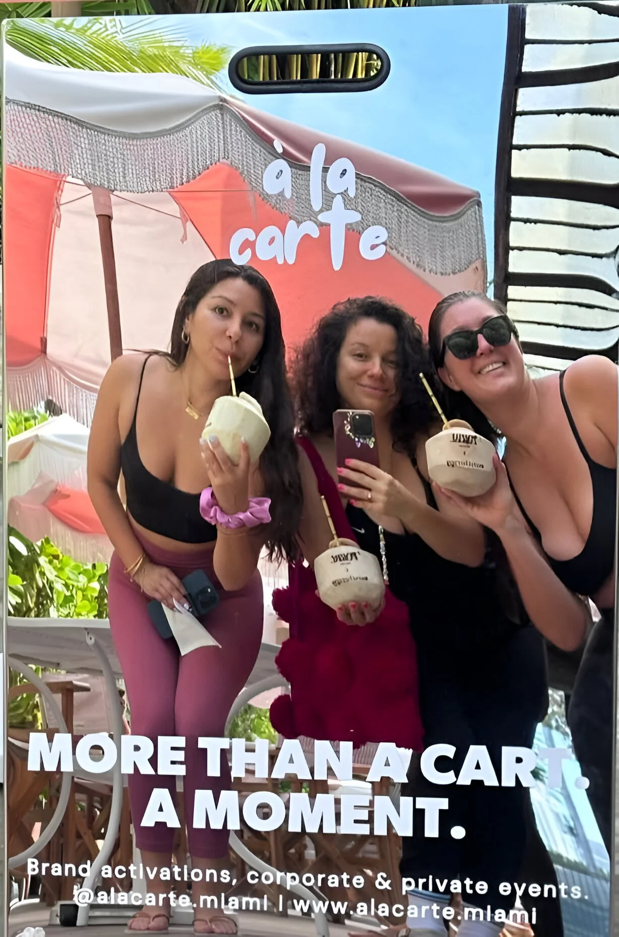 Three women smiling and holding logo coconuts with straws, taking a selfie at an outdoor corporate event.