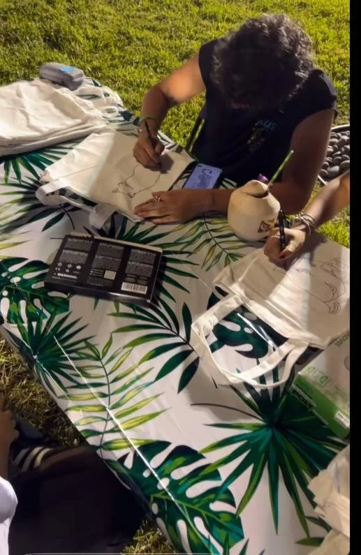 Two children are drawing on tote bags with markers at an outdoor table covered with a tropical leaf-patterned tablecloth. One child has a custom coconut next to them.
