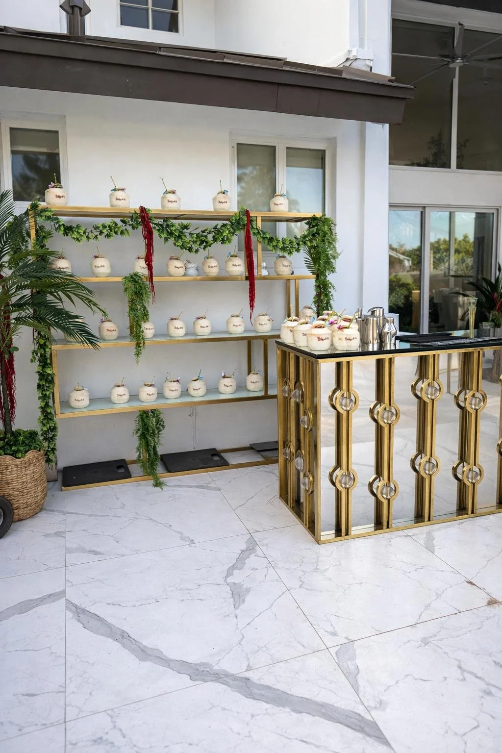 An outdoor bar setup with a gold and glass counter, white marble flooring, and a white wall with a shelving unit decorated with green foliage and strings of red ribbons, holding white coconut drinks with colorful straws and umbrellas, under a shaded 